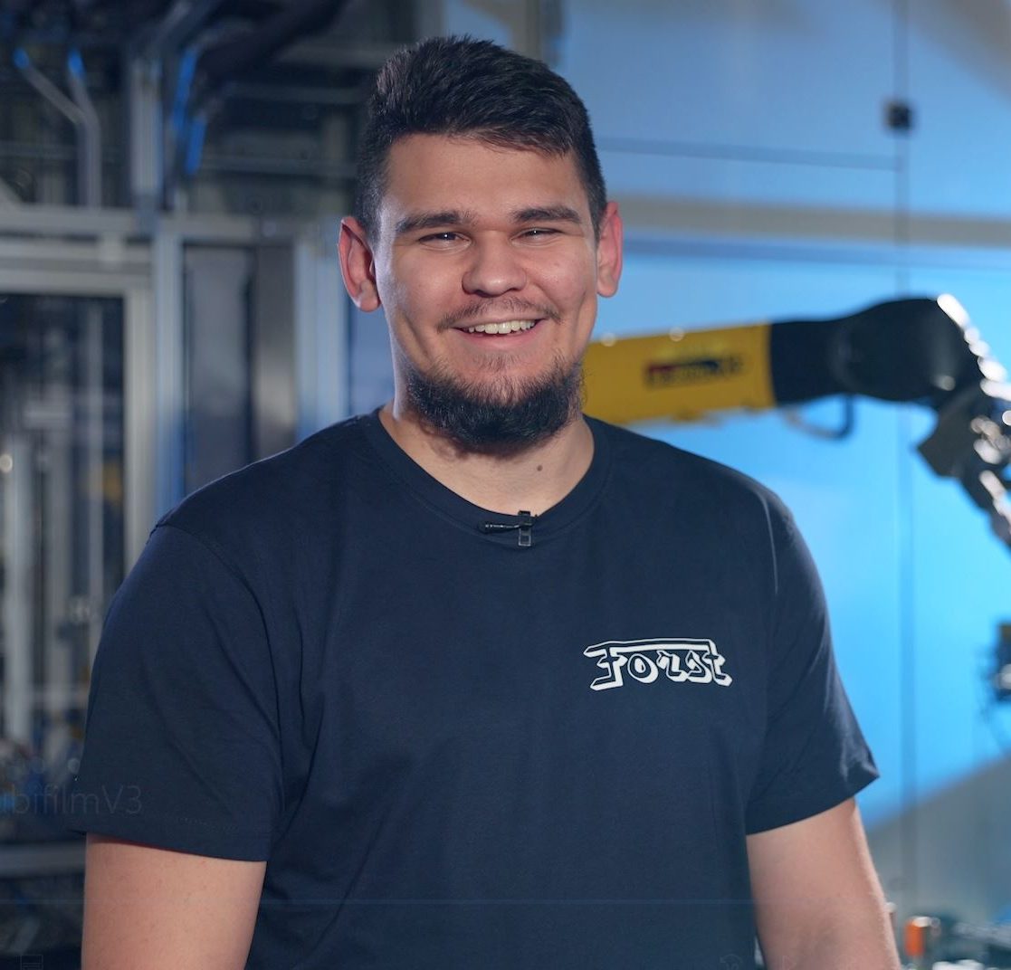 Junger Mann mit dunkelbraunen Haaren und einem Bart, trägt ein dunkelblaues Tshirt mit der Aufschrift "Forst"