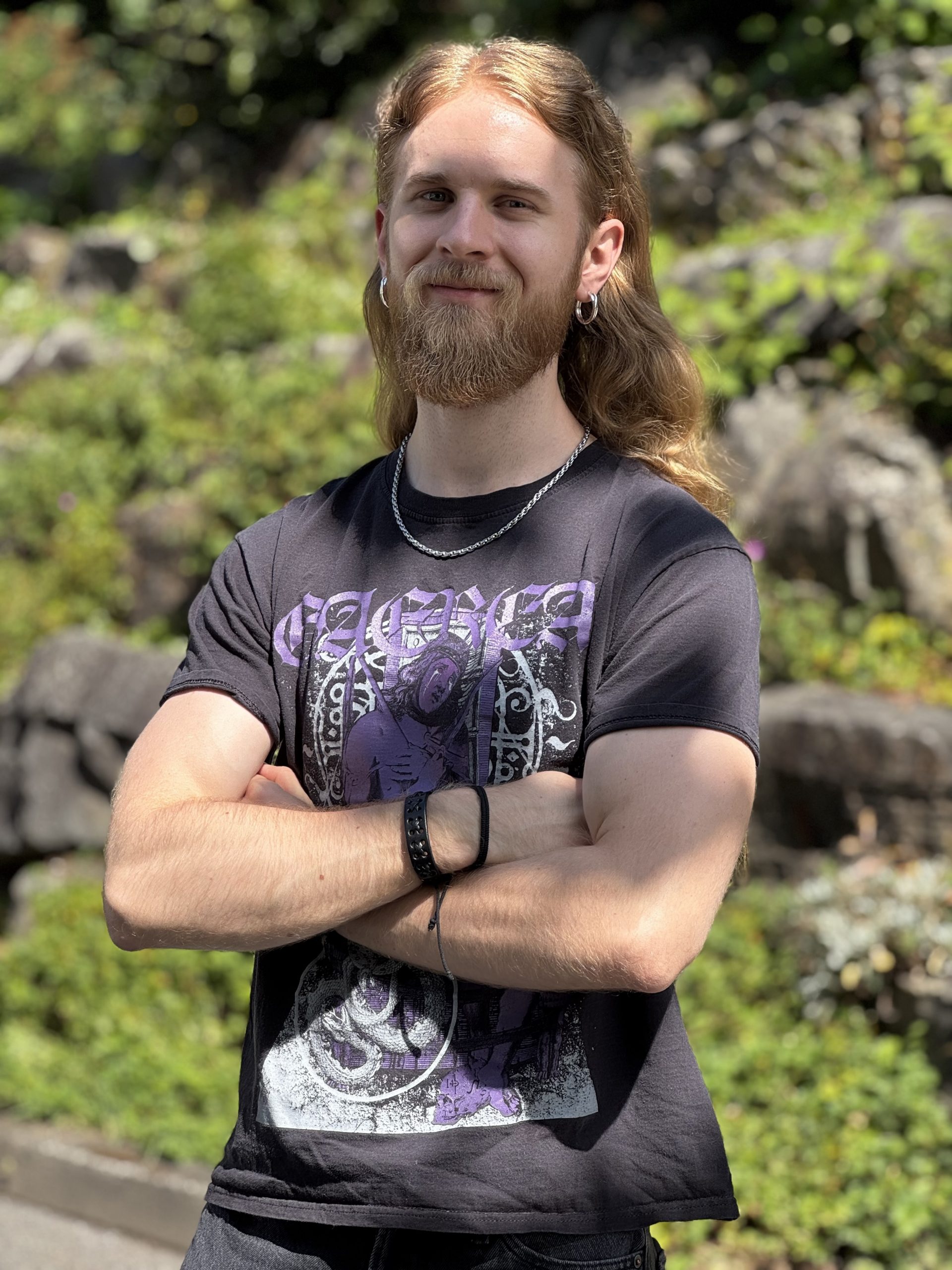 Junger Mann mit Rot-Braunen Haaren und Bart, verschränkt die Arme und trägt ein dunkelgraues T-Shirt mit einem Printzug vorne drauf